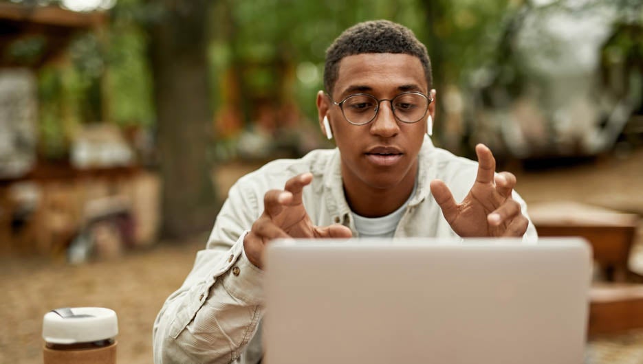 Young african american man taking part in online conference while sitting at wooden table near laptop outdoors and talking into screen. Work in nature concept